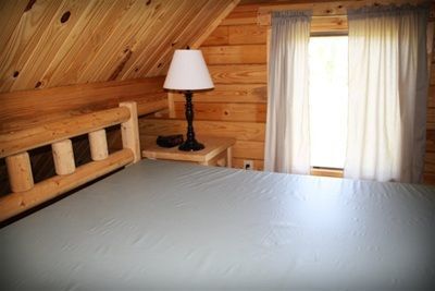 A bedroom in a log cabin with a bed , nightstand , lamp and window.