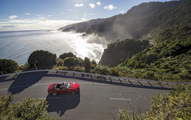 A red convertible is driving down a winding road next to the ocean.