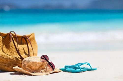 A hat , sunglasses , flip flops and a bag are on the beach.