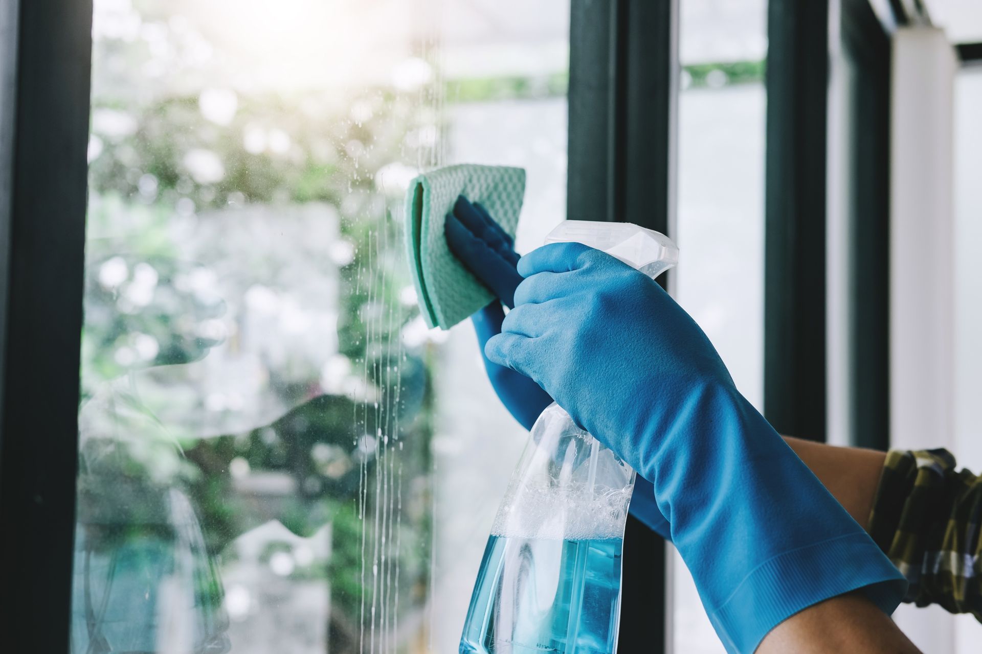 Person wearing blue gloves cleans a window with a sponge and spray bottle.