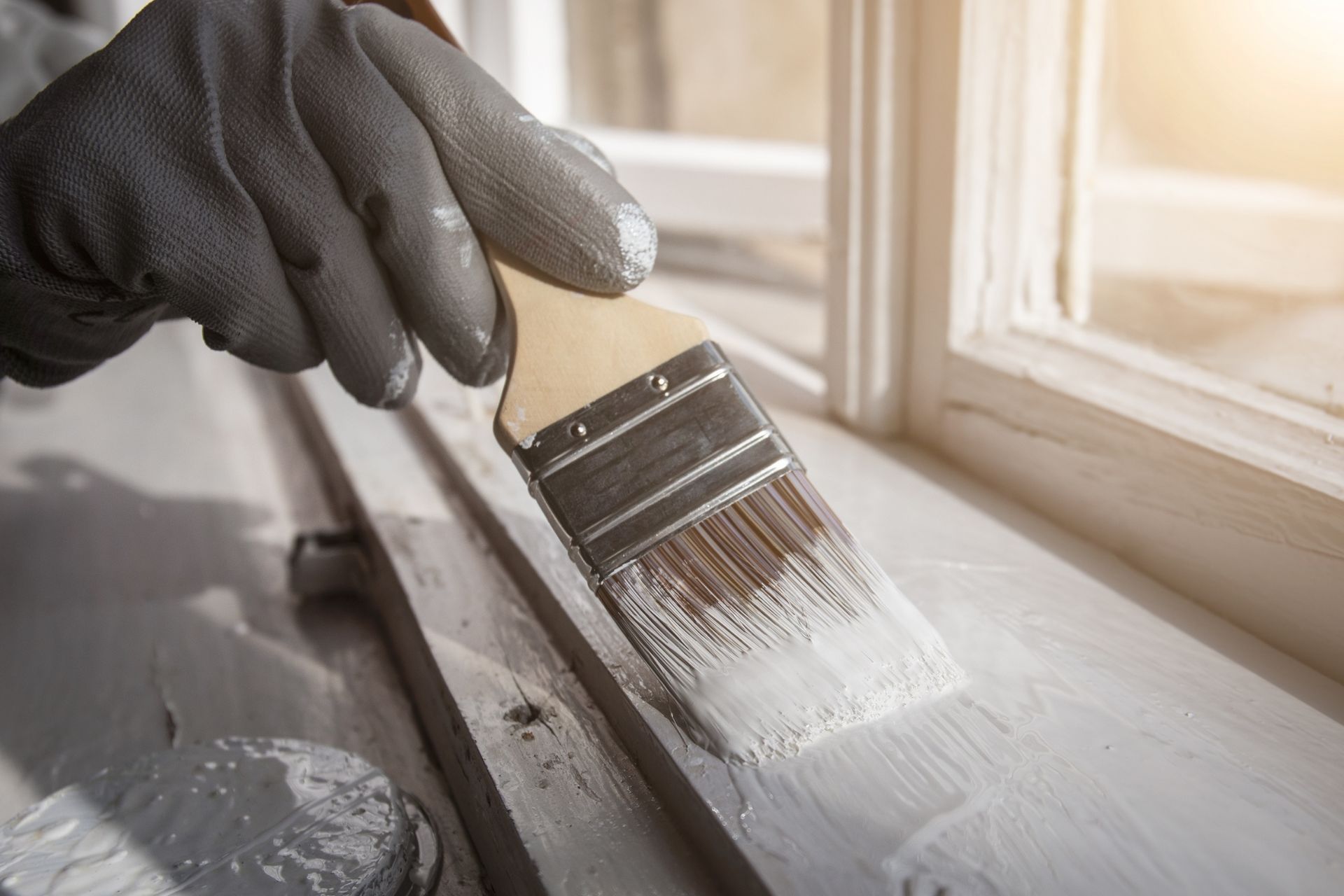 Gloved hand painting white paint on a wooden windowsill near a window. Sunlight illuminates the scene.
