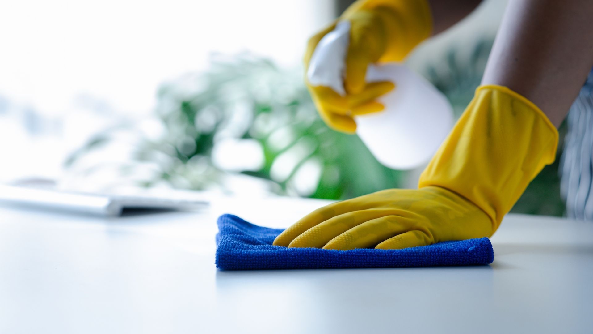 Person wearing yellow gloves sprays and wipes a blue cloth on a white surface, cleaning.