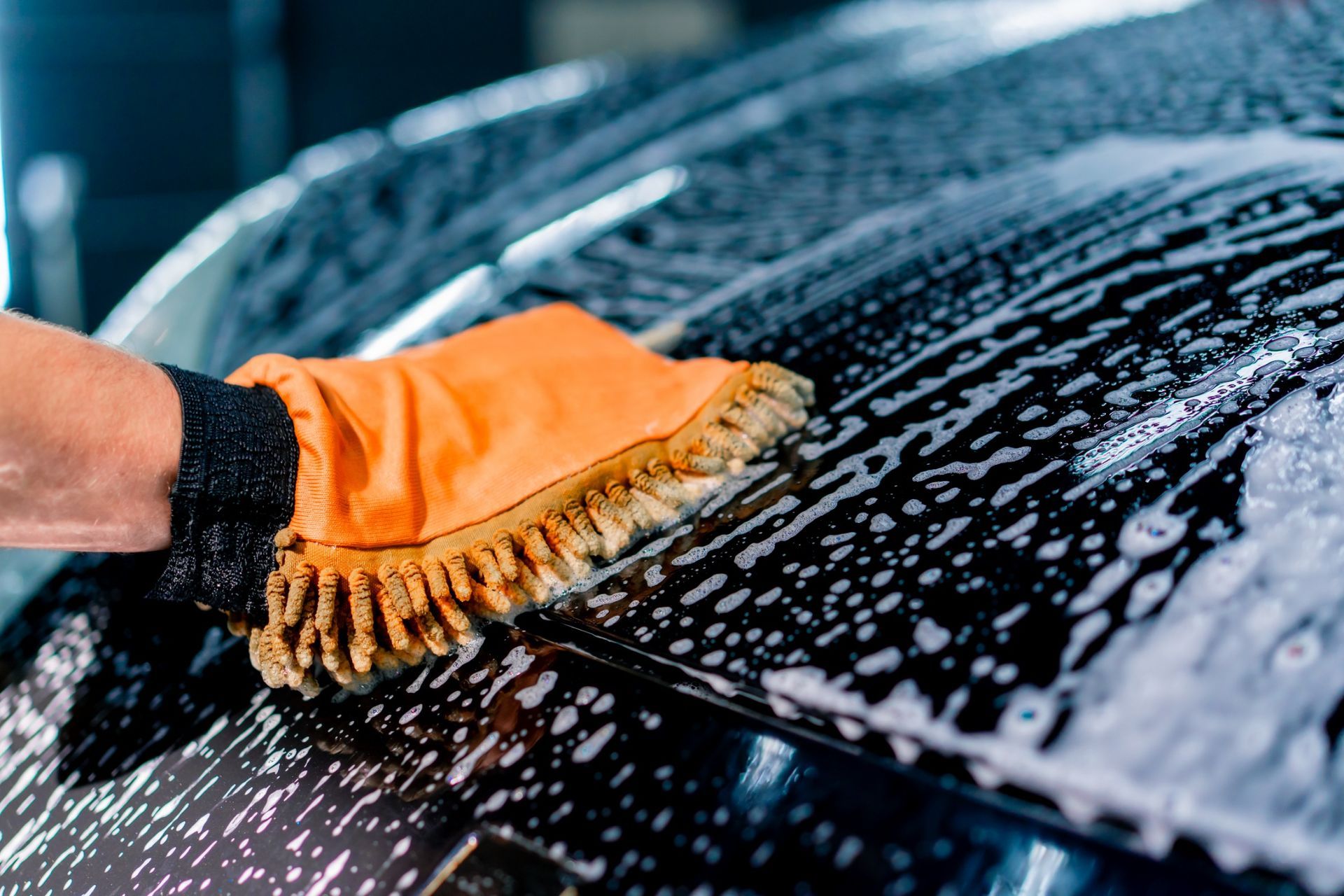 Hand wearing orange mitt washing a black car with suds.