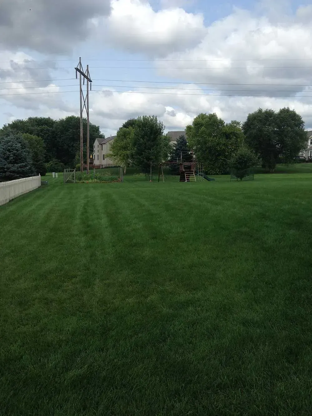 Lush green lawn with a picket fence, trees, and a power line under a cloudy sky.
