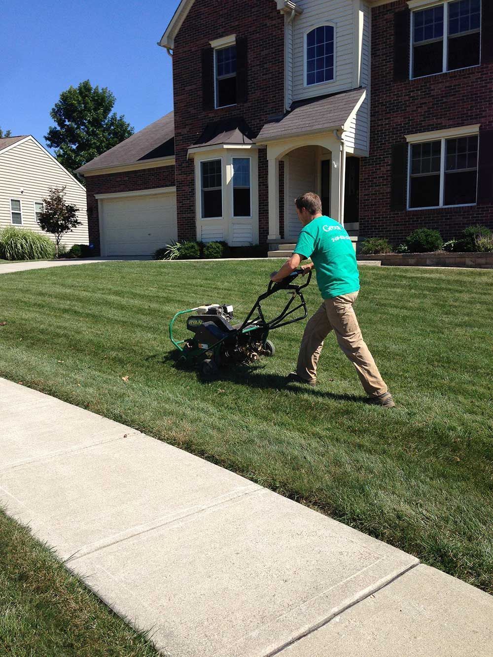 Man mowing a green lawn in front of a brick house on a sunny day.