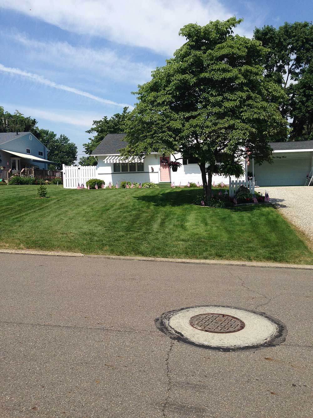 A white house with a large green tree in front. The yard has fresh-cut grass, and a manhole is in the street.