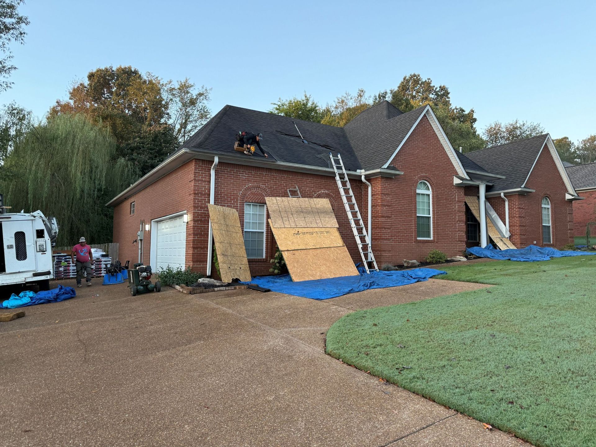 Brick house under construction with blue tarp, plywood-covered windows, and gravel driveway