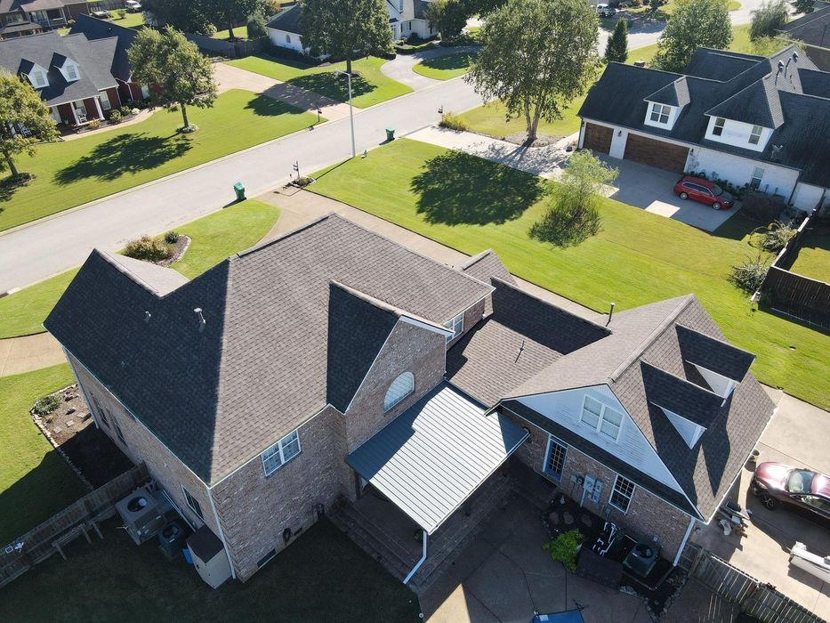 Aerial view of a suburban house with a dark roof, driveway, and tree-lined neighborhood street.