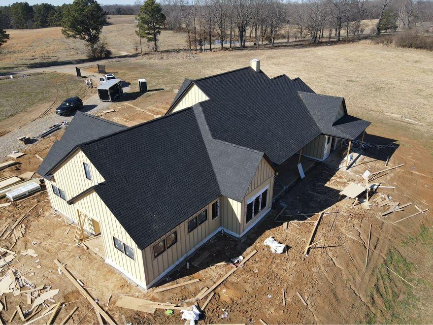 Aerial view of a house under construction with dark roof shingles and unfinished dirt surroundings.