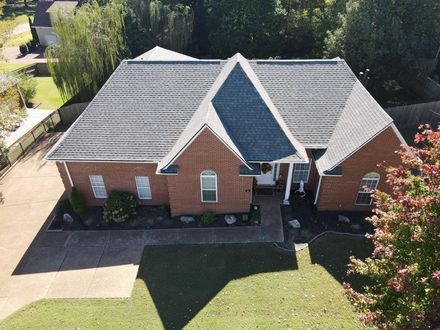 Brick house with gray shingle roof, front porch, and driveway surrounded by green lawn and trees.