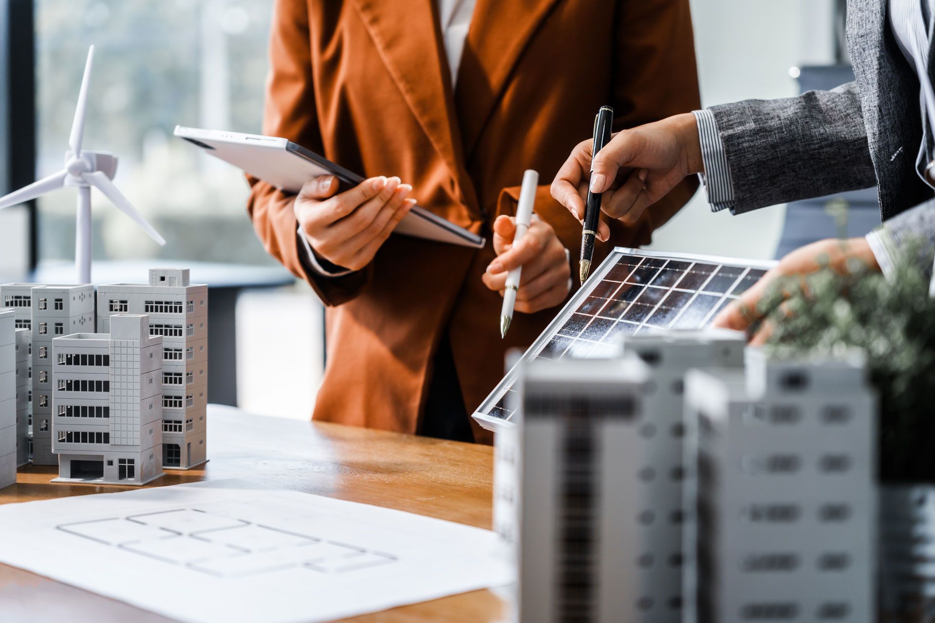 Two people examining solar panel model and building plans, with miniature buildings and wind turbine.