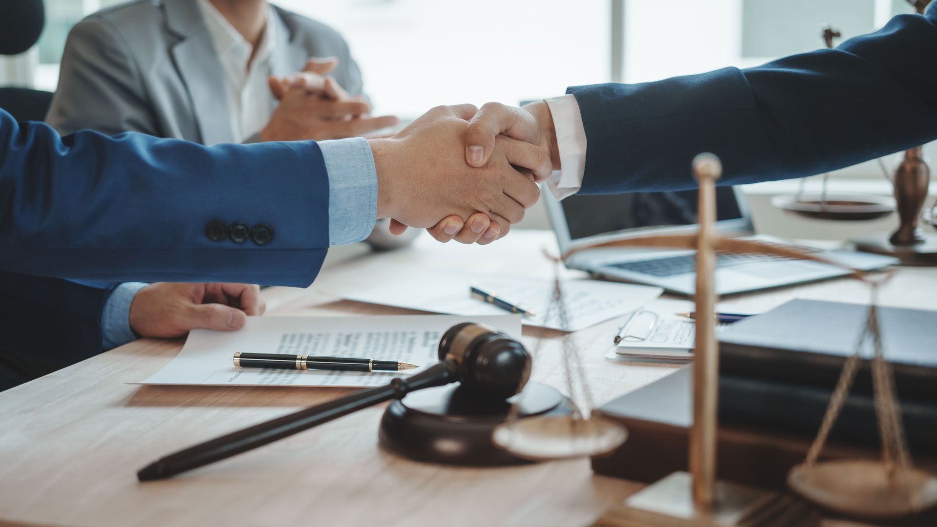 Two men in suits shaking hands at a table with legal documents, scales, and a gavel.