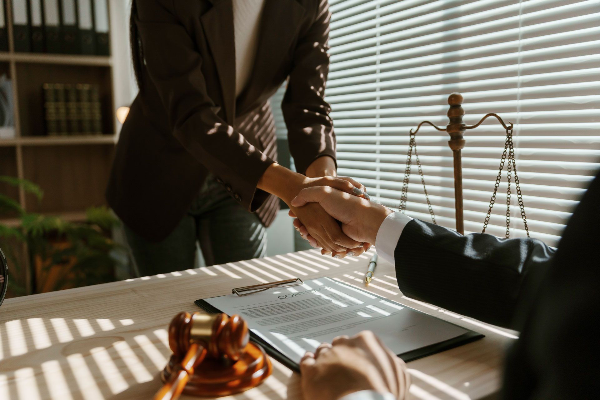 Two people shaking hands over a contract at a desk in a law office. Gavel and scales of justice visible.