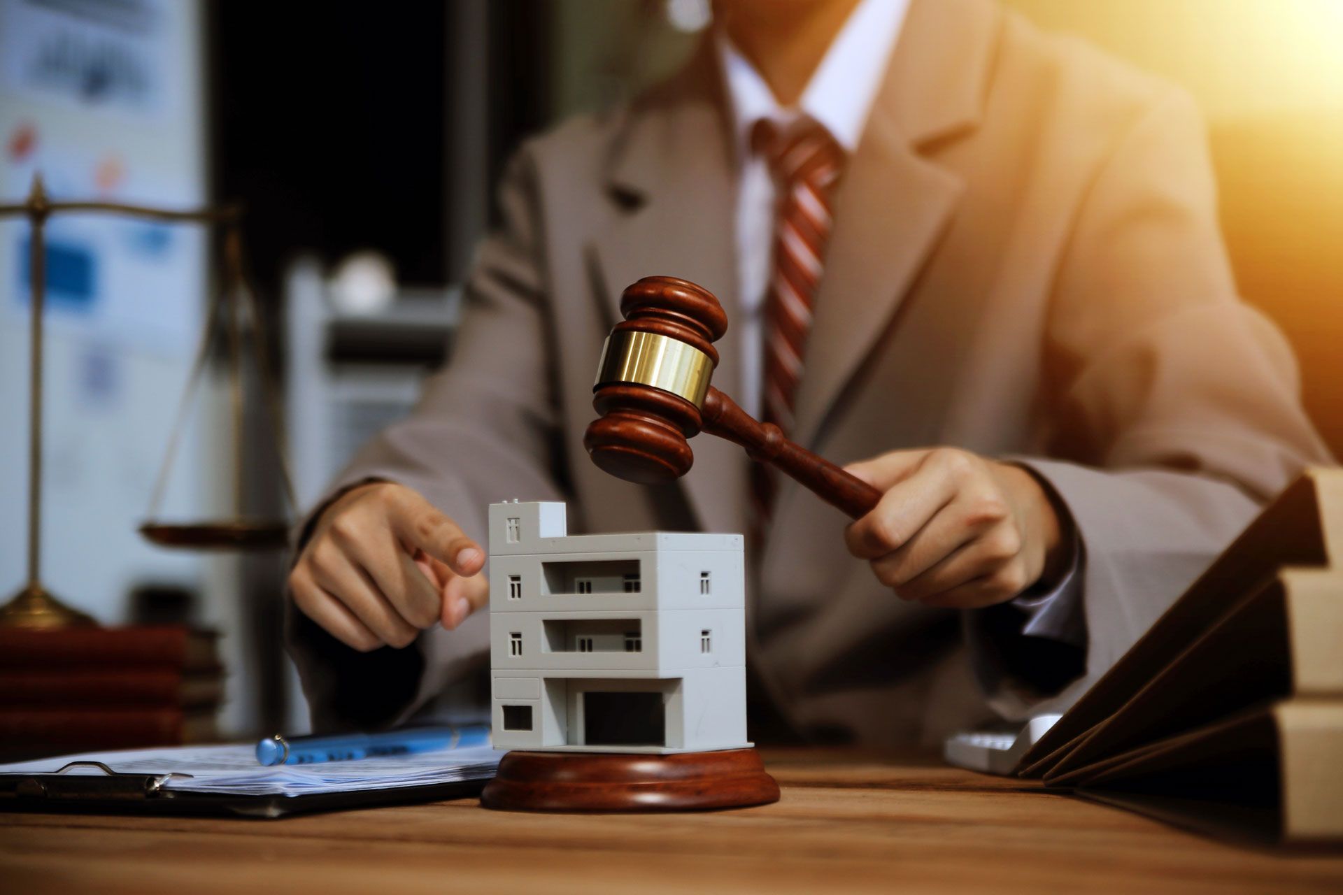 Person in suit holding gavel over a building model on a desk; scales and documents in background.