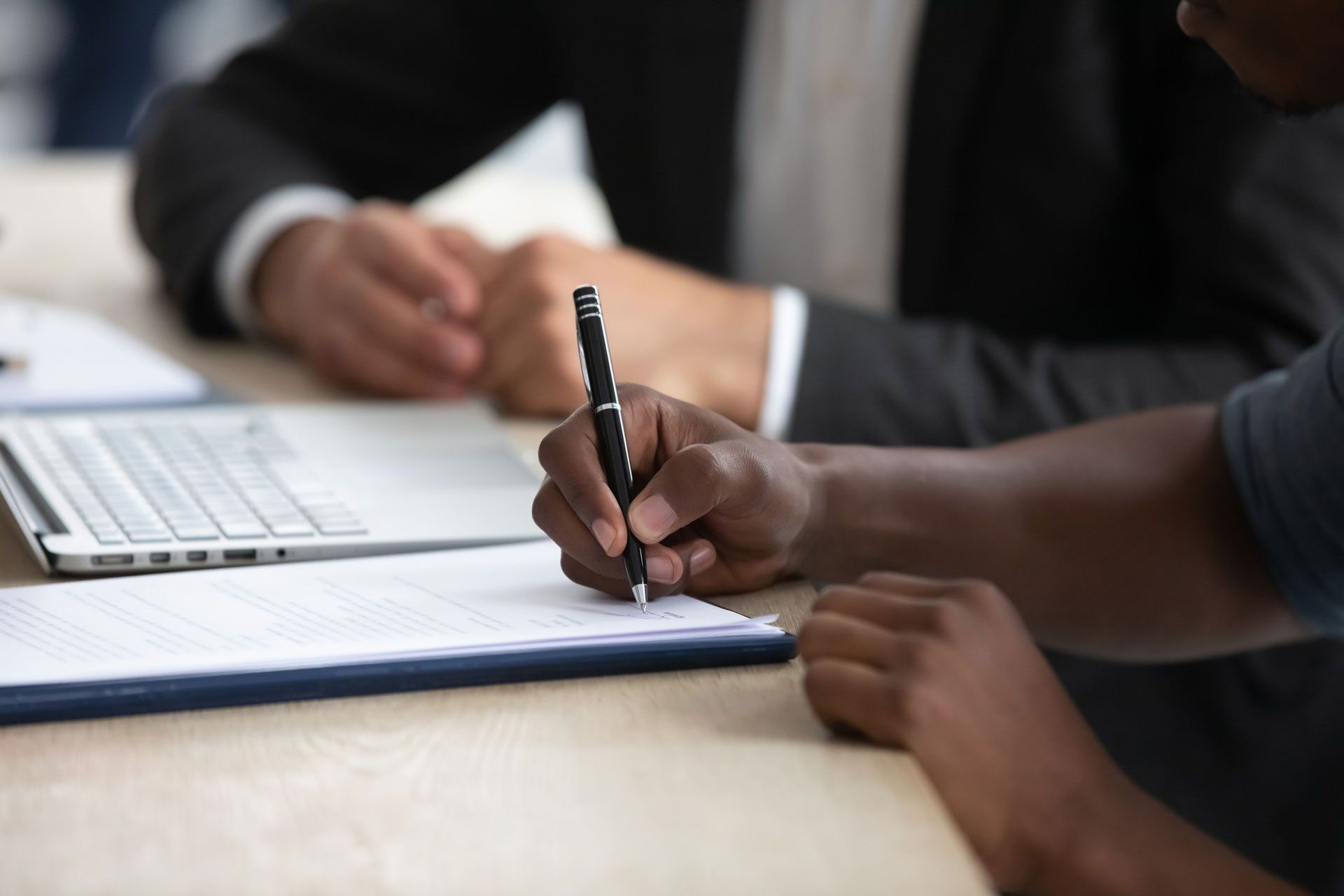 A person signing a document on a clipboard, another person in suit beside a laptop in office setting.