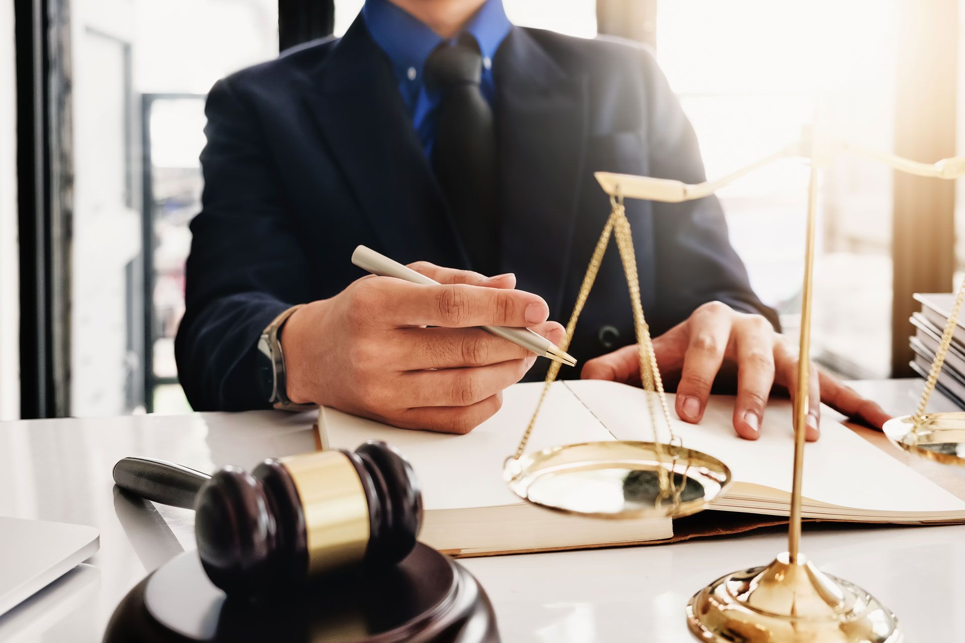 Person in a suit holding a pen, next to a gavel and scales of justice on a desk.