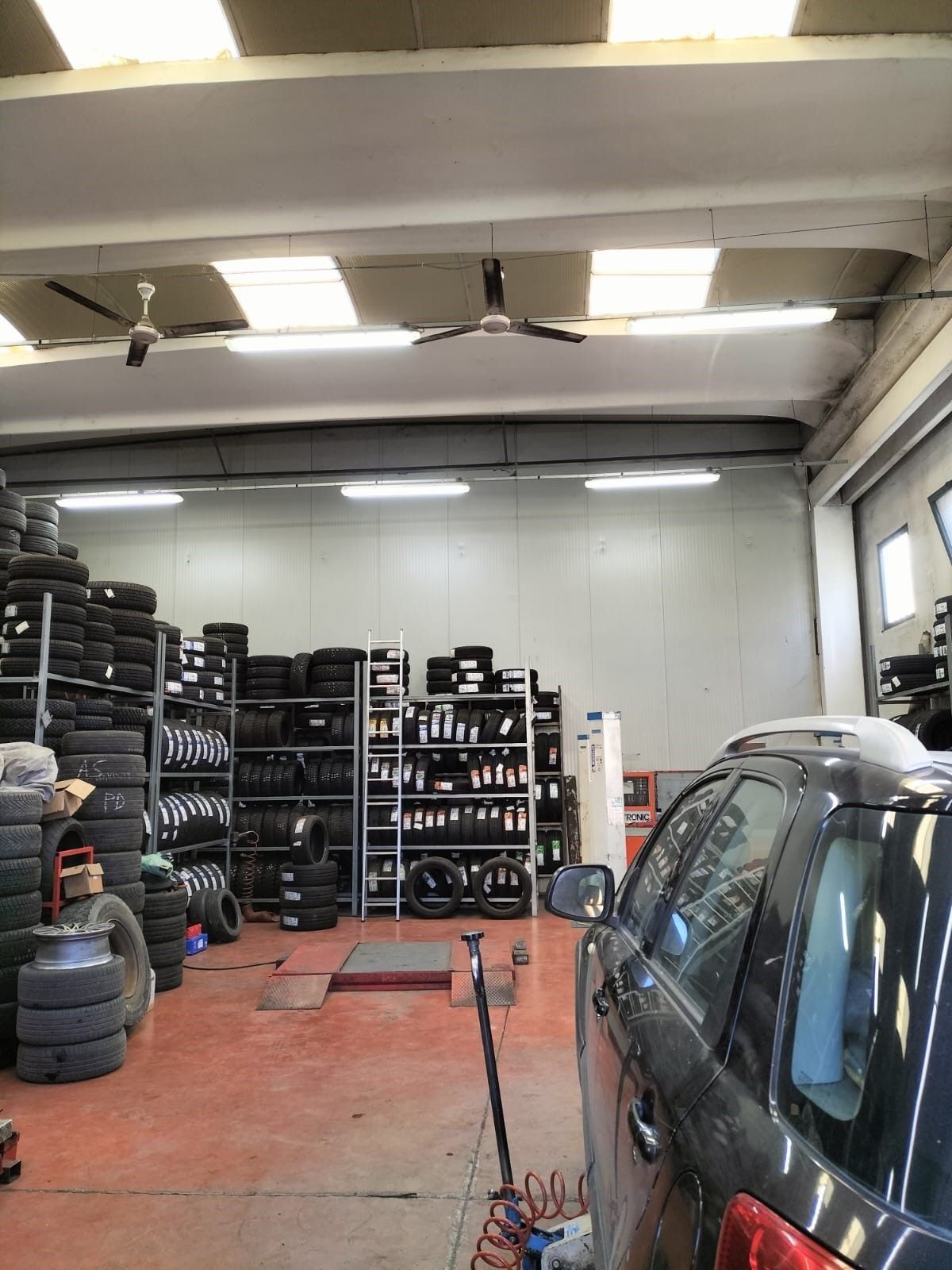 Interno di un gommista con pneumatici impilati, scaffali e un'auto. Ventilatori a soffitto e luci fluorescenti illuminano lo spazio.