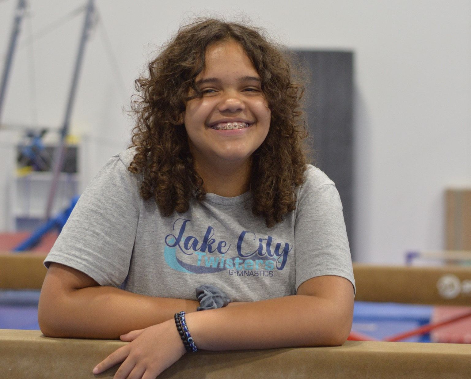 A young girl is leaning on a balance beam and smiling.