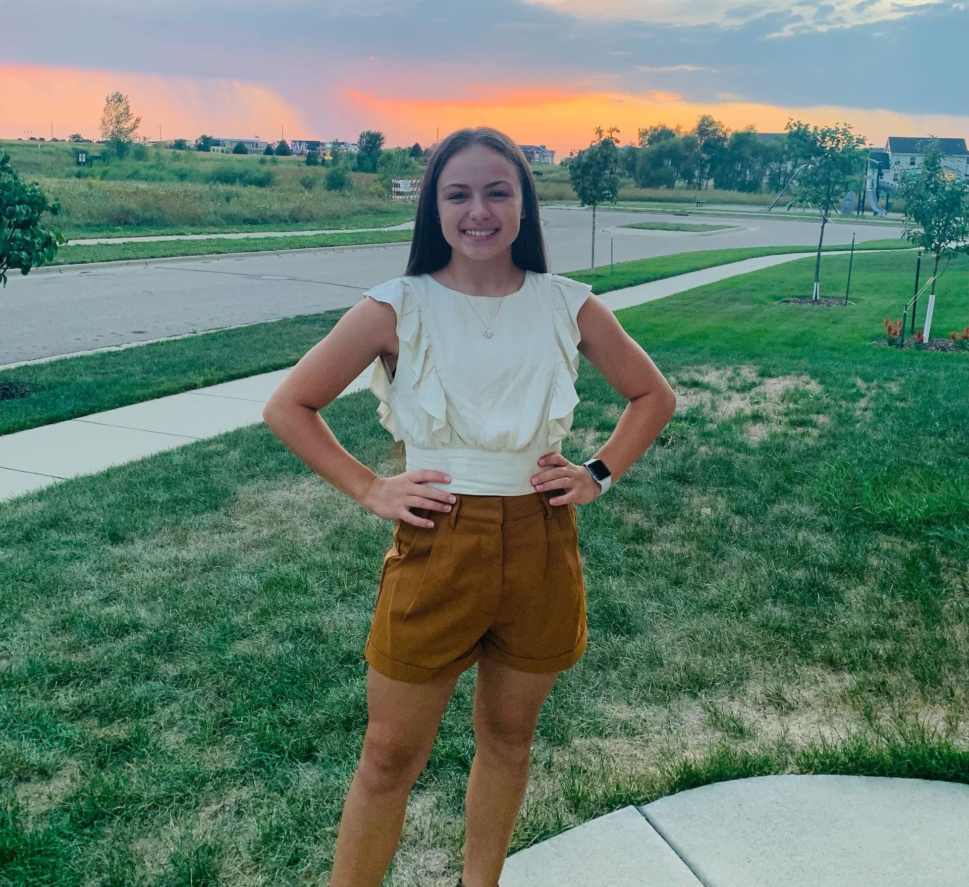 A woman in a white top and brown shorts is standing on a sidewalk with her hands on her hips.