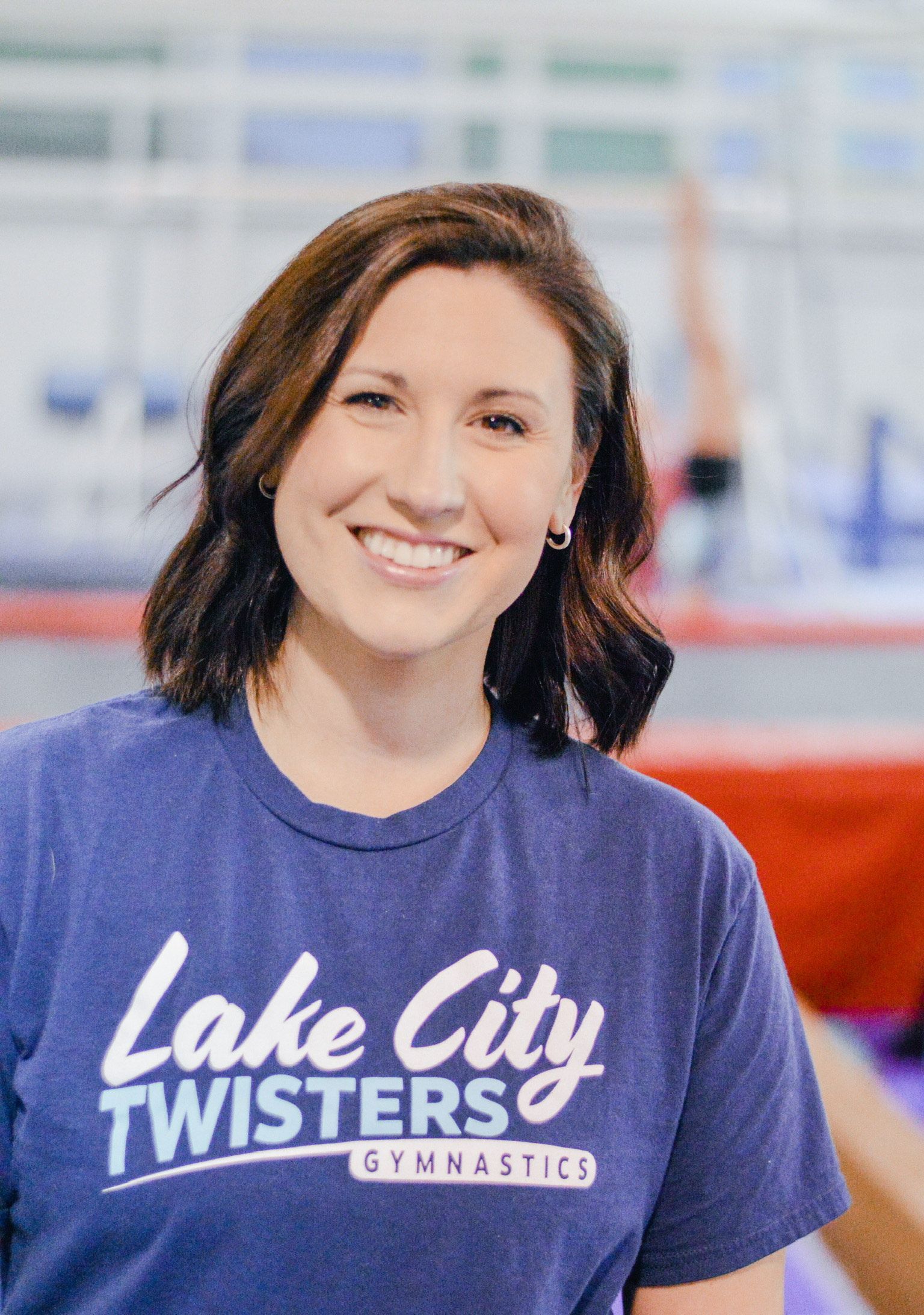 A woman wearing a lake city twisters gymnastics shirt is smiling.