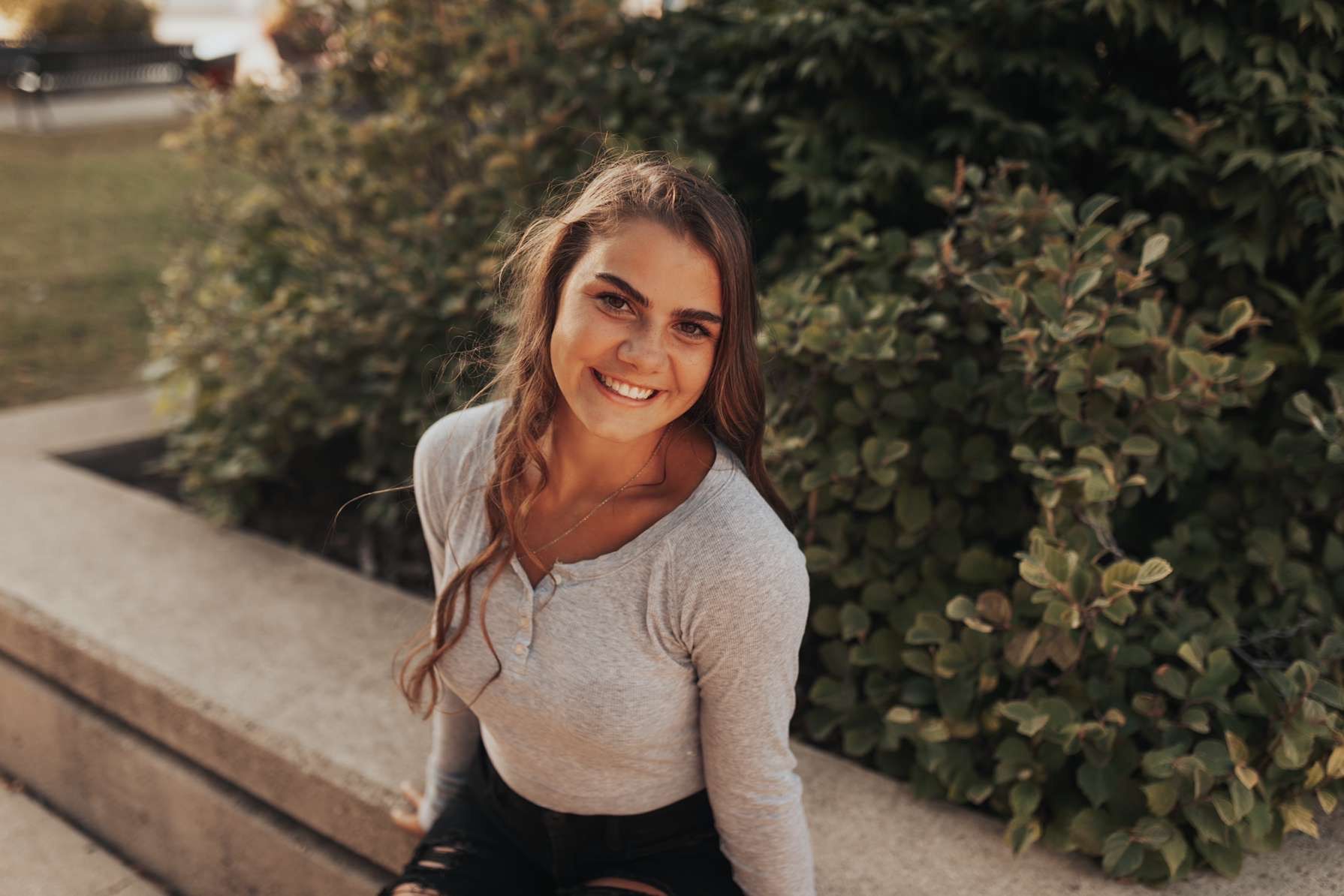 A young woman is sitting on a bench in front of a bush and smiling.