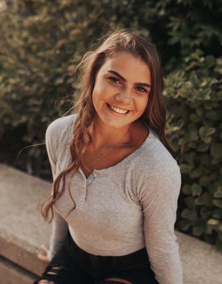 A young woman is sitting on a bench in front of a bush and smiling.