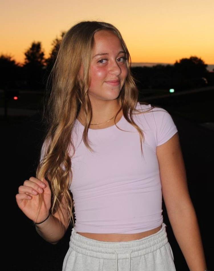 A woman in a white top and brown shorts is standing on a sidewalk with her hands on her hips.