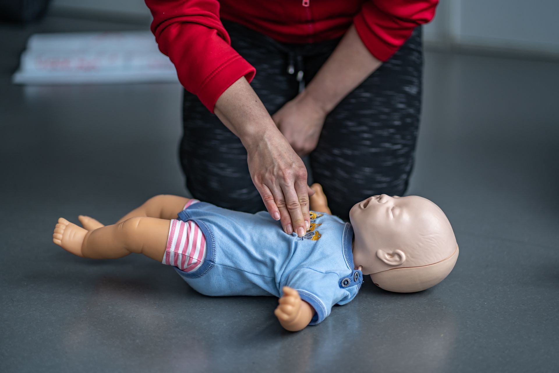 Person practicing infant CPR on a doll, indoors. Hands on doll's chest, legs, blue shirt, red top.