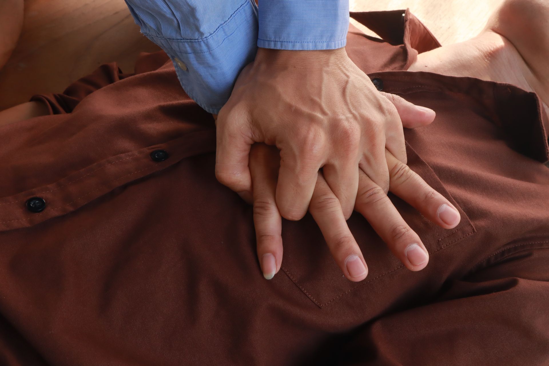 Hands performing CPR on a person's chest, pressing down rhythmically. Brown shirt.