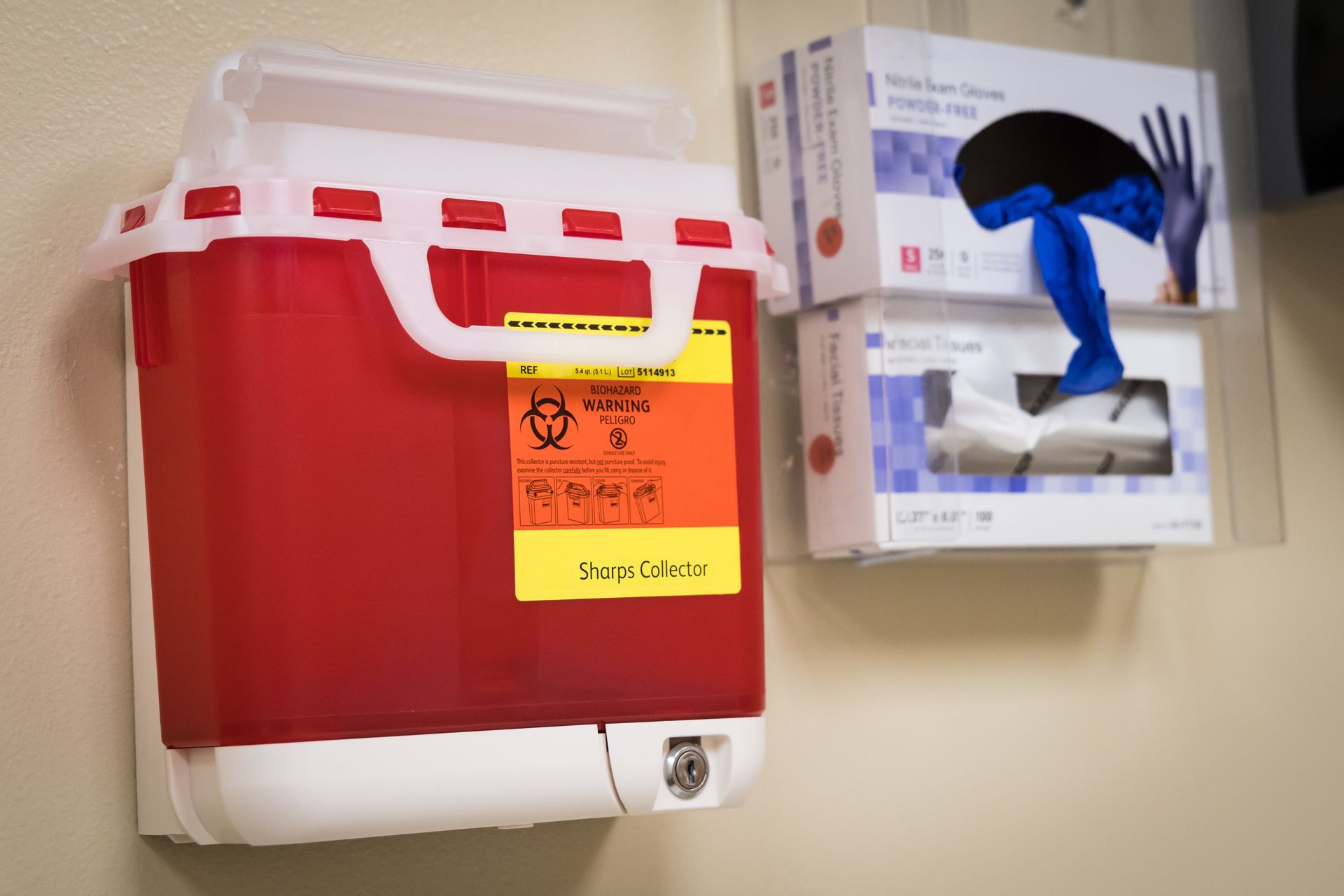 Red sharps container with biohazard label and boxes of gloves on wall.