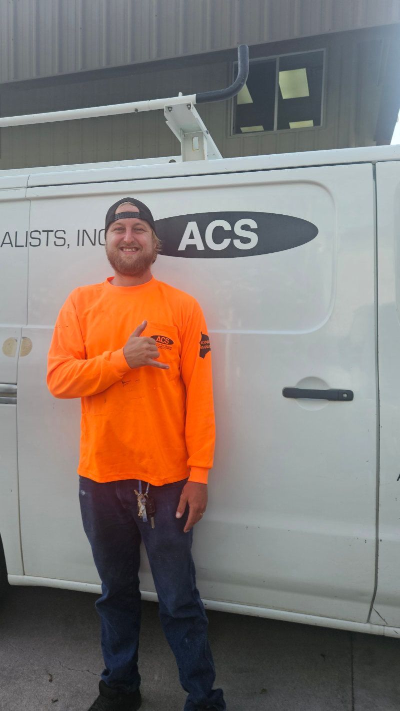A person in a bright orange long-sleeve shirt smiling and giving a shaka sign in front of a white work van labeled ACS.