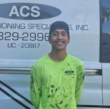 A smiling person in a stained neon green long-sleeved work shirt stands in front of a white van with