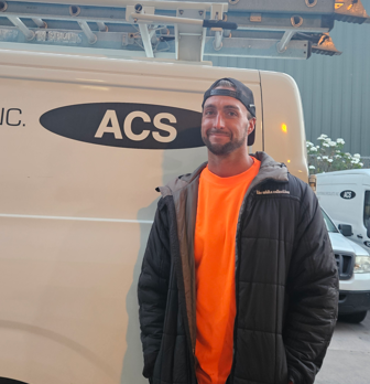 A person in an orange shirt and black jacket stands in front of a white work van with an ACS logo and roof ladder rack.