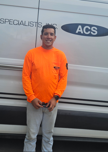 A person in a bright orange long-sleeved shirt and gray pants standing in front of a white work van labeled