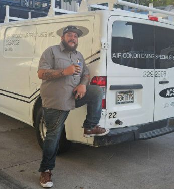 A person with a beard and hat leans against a white Air Conditioning Specialists service van while holding a drink.