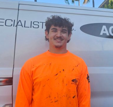 A person wearing a long-sleeved orange shirt stands in front of a white work van with a partial logo.