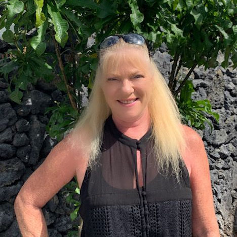 A person with long blonde hair and sunglasses on their head smiles while standing in front of a dark stone wall.