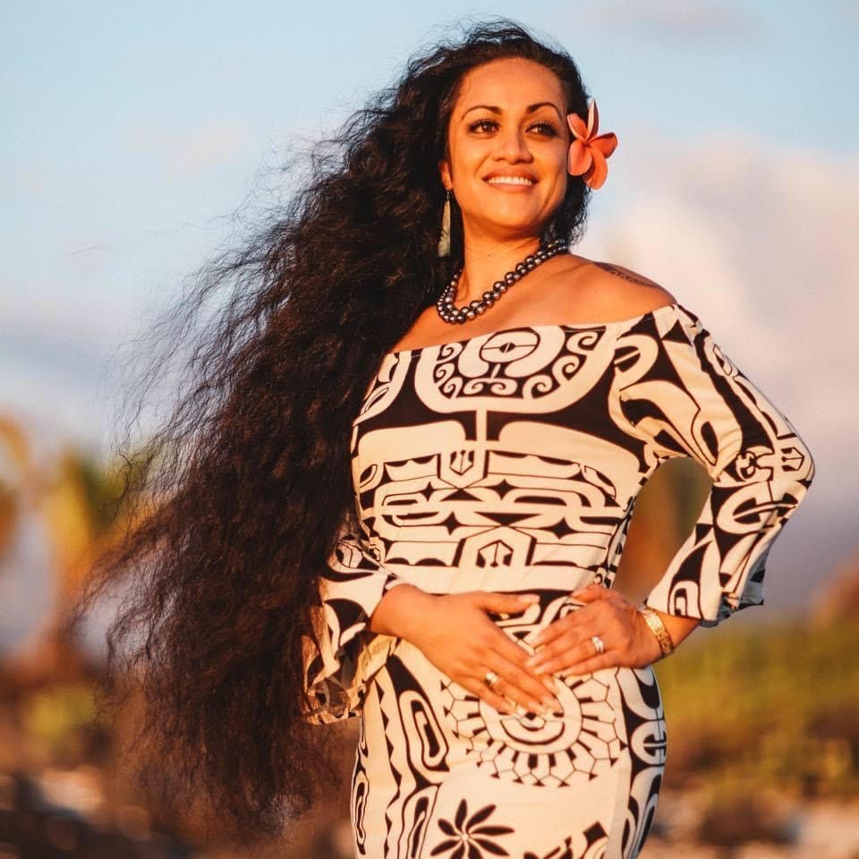 Smiling person with long hair and a flower behind their ear, wearing a patterned dress outdoors in warm, golden light.