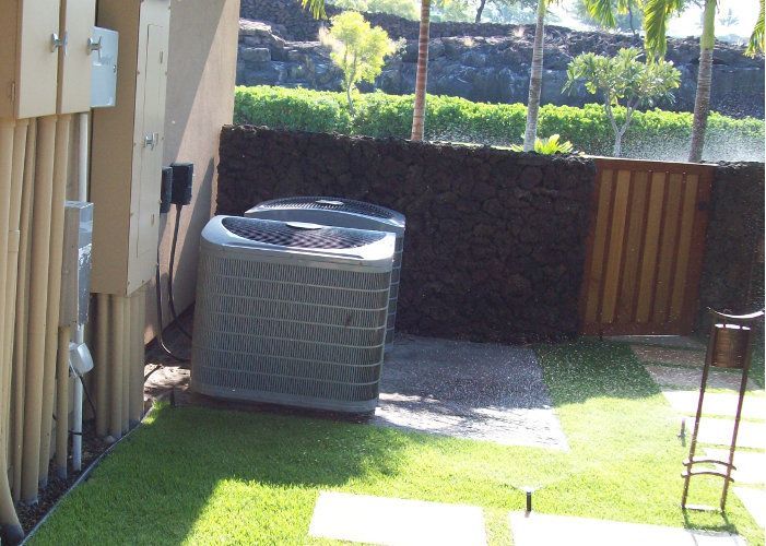 Two gray HVAC units sit on a stone pad outside a building next to a dark rock wall and a wooden gate.