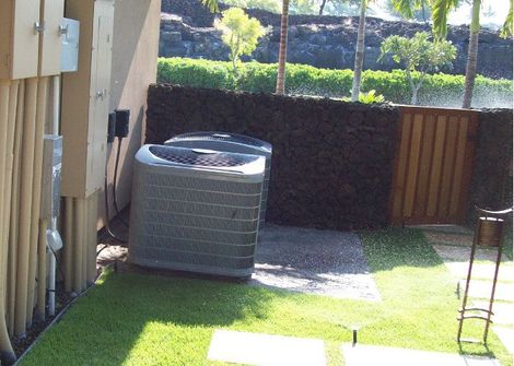 Two gray HVAC units sit on a stone pad outside a building next to a dark rock wall and a wooden gate.