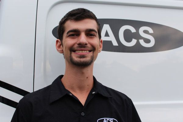 A smiling person wearing a black uniform shirt with an ACS logo, standing in front of a white van with ACS branding.