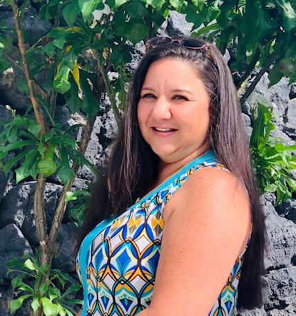 A smiling woman with long brown hair wearing a blue and yellow patterned top stands in front of a tree and rock wall.