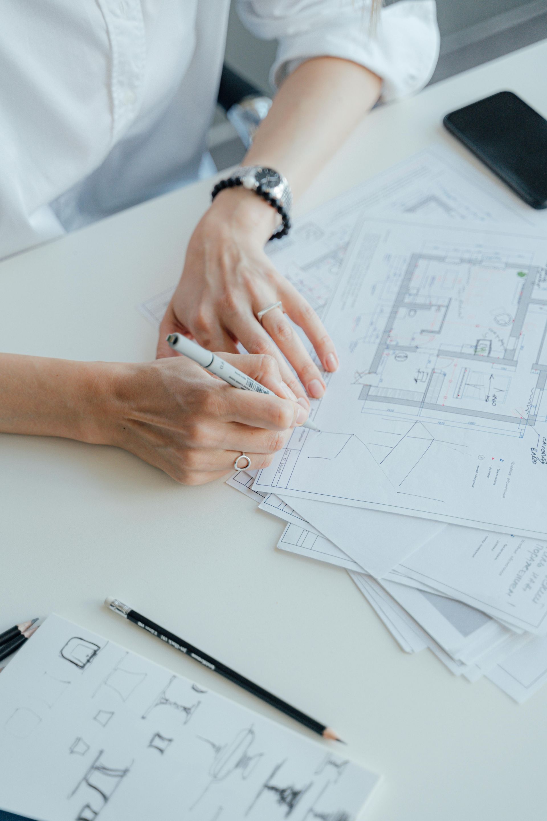 A person wearing a white shirt and watch writes with a pen on architectural floor plans on a desk.