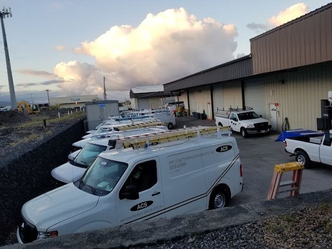 A row of white service vans with ladders parked outside a large warehouse building under a cloudy sky.