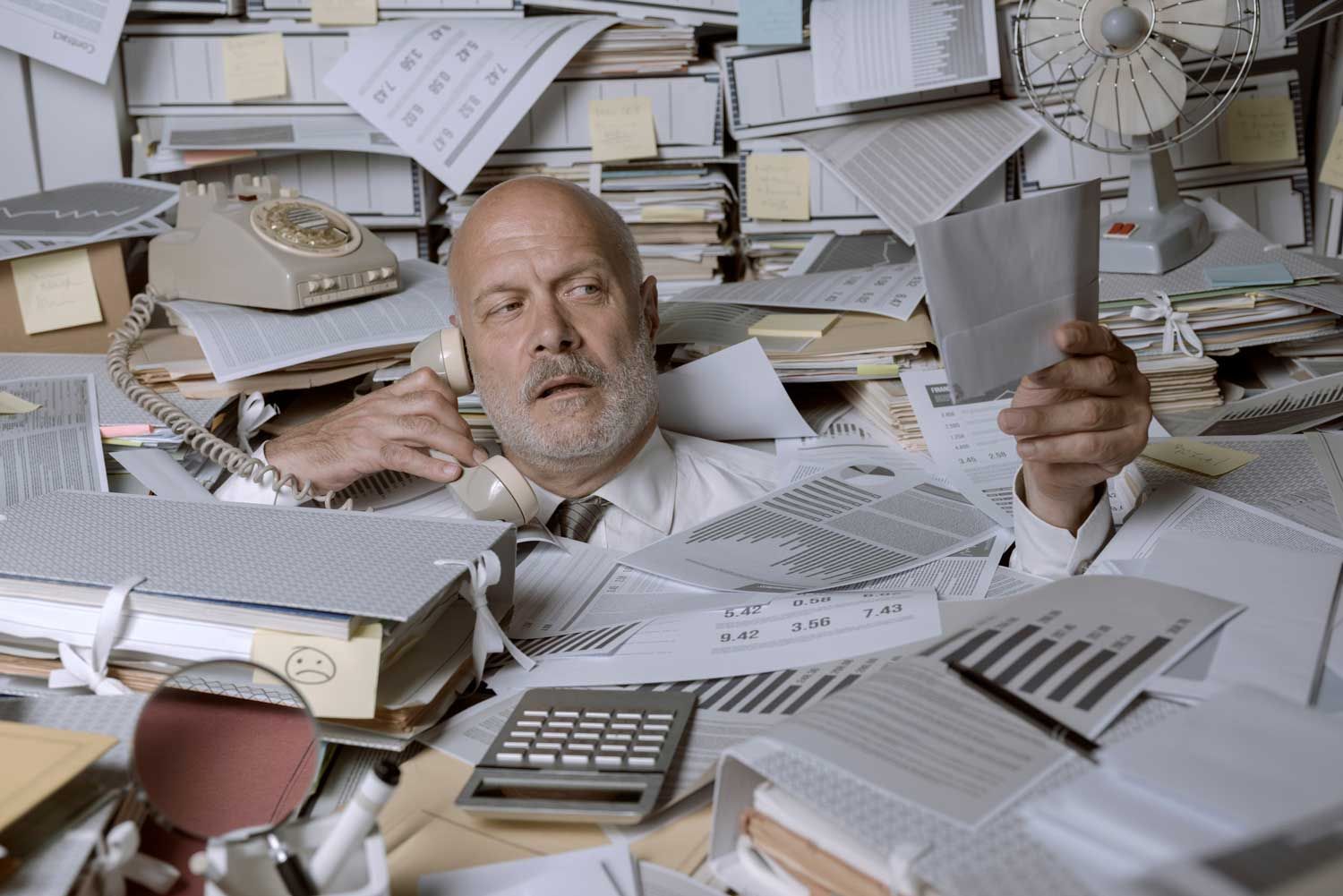 A businessman buried in paperwork, on the phone, looking overwhelmed at his desk.