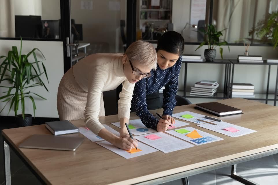Two women in an office, reviewing documents together, one pointing and writing on a paper.
