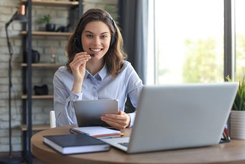Woman with headset smiles, holding tablet, working at a laptop, in a home office setting.