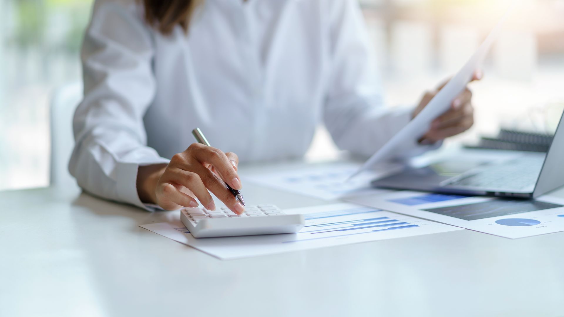Woman calculating finances at a desk with papers, graphs, calculator, and laptop.