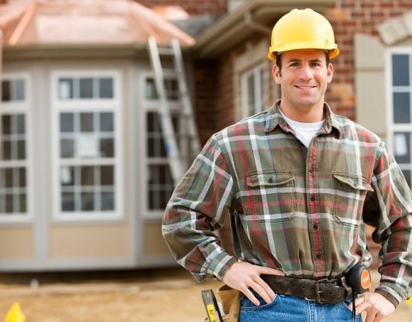 Construction worker in plaid shirt and yellow hard hat smiles in front of a building under construction.