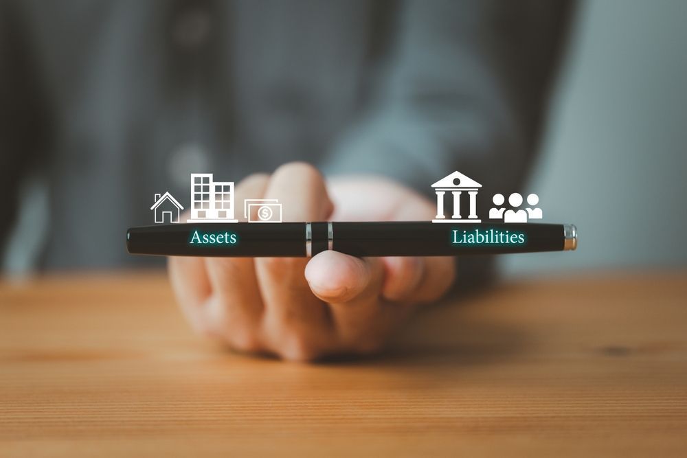 A businessperson balancing icons labeled assets and liabilities on a wooden block